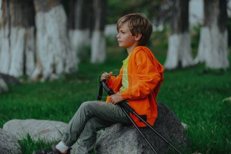 Foto de Niño excursionista con bastones de trekking sentado en una roca y mirando hacia otro lado - Imagen libre de derechos