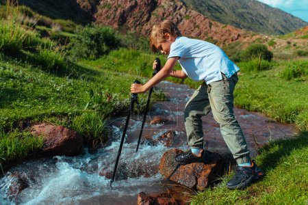 Foto de Niño con bastones de trekking comprueba la profundidad del arroyo de montaña - Imagen libre de derechos