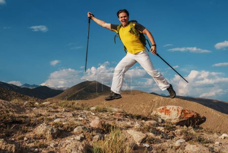 Foto de Feliz joven excursionista masculino con bastones de trekking y mochila saltando sobre el fondo de las montañas - Imagen libre de derechos