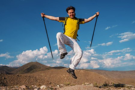Foto de Feliz joven excursionista masculino con bastones de trekking y mochila saltando sobre el fondo de las montañas - Imagen libre de derechos