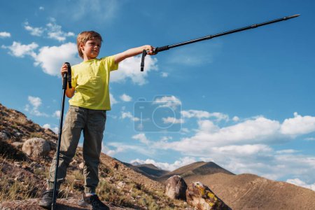 Photo for Boy hiker with trekking poles stands on top of a mountain and points into the distance - Royalty Free Image
