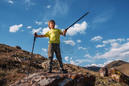 Téléchargez les photos : Garçon randonneur avec bâtons de trekking se dresse au sommet d'une montagne et des points dans la distance - en image libre de droit
