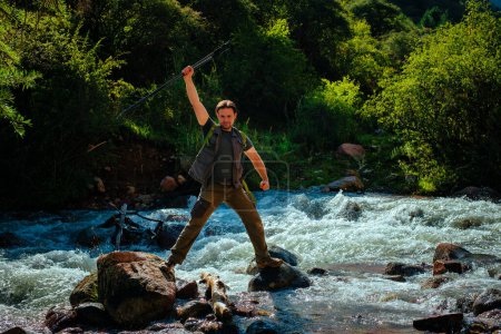 Téléchargez les photos : Heureux randonneur avec des bâtons de trekking se dresse au milieu d'une rivière de montagne - en image libre de droit