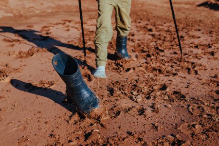 Téléchargez les photos : Garçon randonneur avec bâtons de trekking a perdu sa botte en caoutchouc dans la boue, se concentrer sur la botte - en image libre de droit