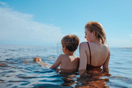 Photo for Mother and son sitting on seashore in water and looking away - Royalty Free Image