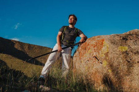 Téléchargez les photos : Randonneur fatigué avec sac à dos et bâtons de trekking repose par un énorme rocher dans les montagnes - en image libre de droit