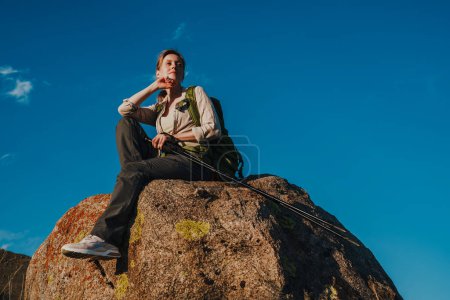 Photo for Young woman tourist with backpack and trekking poles sits on a boulder and looking away - Royalty Free Image