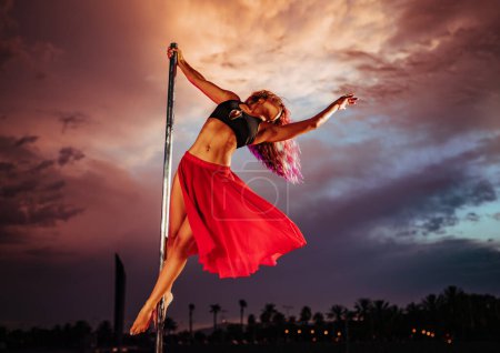 Photo for Young beautiful woman pole dancing on twilight sky and city silhouettes background - Royalty Free Image
