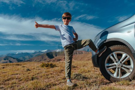 Foto de Niño feliz en gafas de sol se para junto al coche en las montañas y da un pulgar hacia arriba, concepto de viaje - Imagen libre de derechos