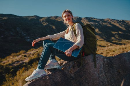 Téléchargez les photos : Jeune femme touriste avec sac à dos et ordinateur portable se trouve au sommet de la montagne - en image libre de droit
