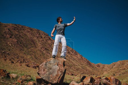 Photo for Handsome young man stands on big stone on mountains background and looks at his hand - Royalty Free Image