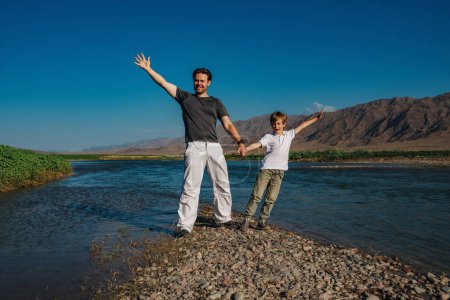 Foto de Feliz padre e hijo de pie en la orilla del lago de montaña cogidos de la mano - Imagen libre de derechos
