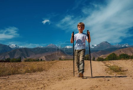 Foto de Niño viajero con bastones de trekking caminando por la carretera de las montañas en verano - Imagen libre de derechos