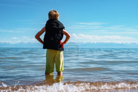 Foto de Niño en chaleco y pantalones cortos se encuentra en el lago y mira a la distancia - Imagen libre de derechos