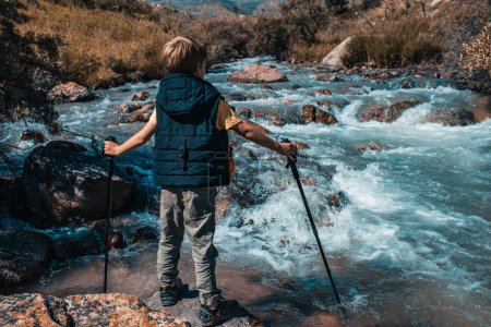 Téléchargez les photos : Garçon randonneur avec bâtons de randonnée debout sur la pierre à la rivière de montagne en automne - en image libre de droit
