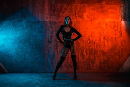 Téléchargez les photos : Jeune danseuse brune en costume noir pose sur fond mural en lumière rouge et bleue - en image libre de droit