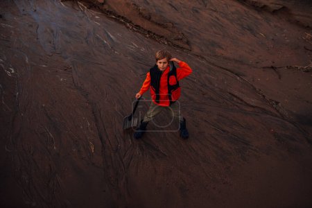 Téléchargez les photos : Garçon avec pelle debout dans un lit de rivière après de fortes pluies - en image libre de droit