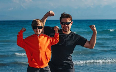 Foto de Padre e hijo en gafas de sol mostrando sus músculos en la playa - Imagen libre de derechos