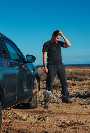 Téléchargez les photos : Homme frustré avec pelle se tient près de sa voiture coincé dans le sable - en image libre de droit