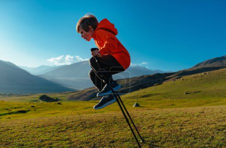 Téléchargez les photos : Joyeux garçon randonneur saute avec des bâtons de trekking dans les montagnes - en image libre de droit