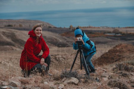 Photo for Boy taking pictures with camera on tripod with his mother in the mountains - Royalty Free Image