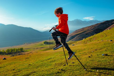 Téléchargez les photos : Joyeux garçon randonneur saute avec des bâtons de trekking dans les montagnes - en image libre de droit