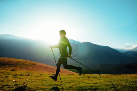 Téléchargez les photos : Jeune homme athlétique avec des bâtons de trekking courant sur fond de montagnes au coucher du soleil - en image libre de droit