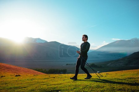 Téléchargez les photos : Jeune homme athlétique avec des bâtons de trekking marchant sur fond de montagnes au coucher du soleil - en image libre de droit