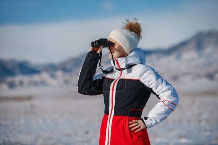 Photo for Young woman tourist looking through binoculars on mountains background in winter day - Royalty Free Image