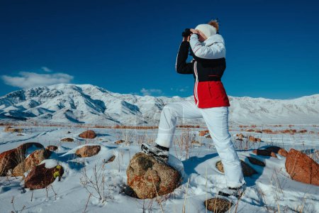 Téléchargez les photos : Jeune femme touriste regardant à travers des jumelles sur fond de montagnes dans la journée d'hiver - en image libre de droit