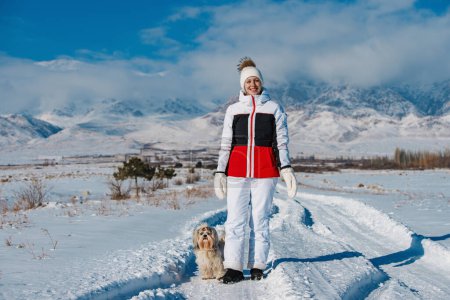 Foto de Feliz joven turista caminando con shih tzu perro en las montañas de fondo en el día de invierno - Imagen libre de derechos