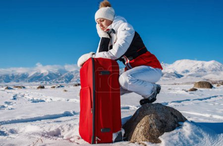 Téléchargez les photos : Triste touriste femme assise sur une pierre avec valise rouge sur fond de montagnes d'hiver - en image libre de droit