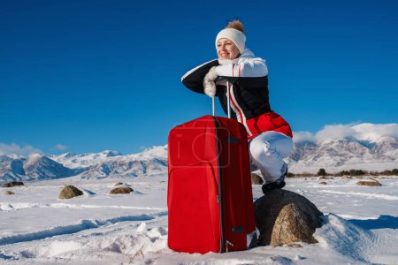 Foto de Mujer joven turista se sienta en una piedra con maleta roja en el fondo de las montañas de invierno - Imagen libre de derechos