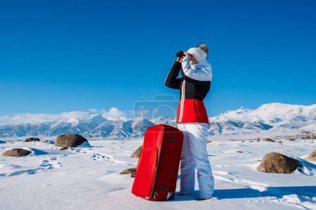 Téléchargez les photos : Jeune femme touriste avec valise rouge regardant à travers des jumelles sur fond de montagnes d'hiver - en image libre de droit
