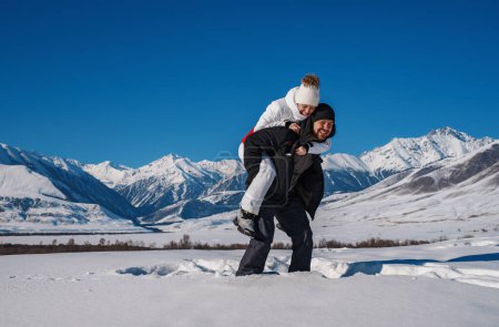 Photo for Happy young couple of tourists having fun in the snowy mountains in winter, man carries woman on his back - Royalty Free Image
