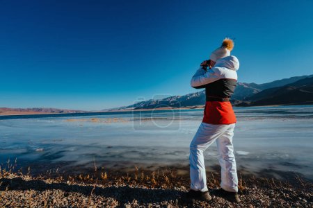 Téléchargez les photos : Jeune femme touriste debout sur la rive du lac et regardant à travers des jumelles en hiver - en image libre de droit