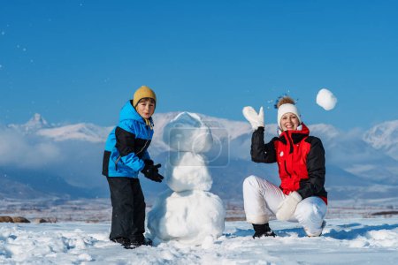 Téléchargez les photos : Joyeux maman et fils faisant bonhomme de neige et jouer boules de neige dans les montagnes - en image libre de droit