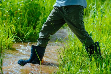 Téléchargez les photos : Garçon en bottes en caoutchouc marchant sur le petit ruisseau sur le champ d'été - en image libre de droit
