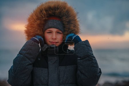 Foto de Retrato de niño en chaqueta de invierno con capucha en la orilla del mar en tiempo tormentoso - Imagen libre de derechos