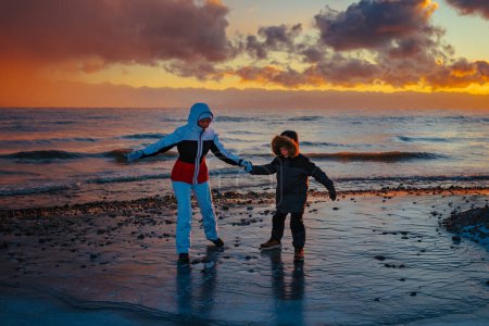 Photo for Mother and son in winter suits standing on slippery ice by lake shore at sunset - Royalty Free Image