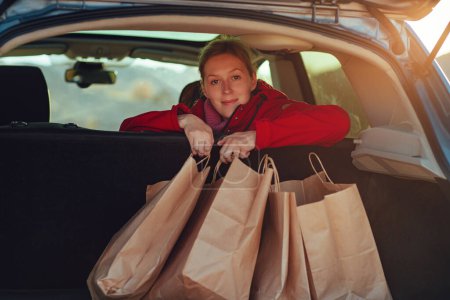 Téléchargez les photos : Heureuse jeune femme tient des sacs à provisions en papier dans la voiture à la lumière du coucher du soleil - en image libre de droit