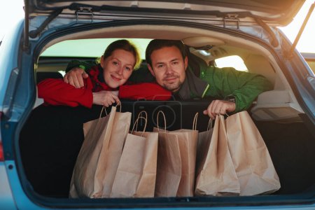 Foto de Feliz pareja joven sostiene bolsas de papel en el coche al atardecer - Imagen libre de derechos
