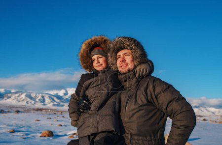 Foto de Retrato de feliz padre e hijo en chaquetas de invierno con capucha sobre fondo de montaña - Imagen libre de derechos