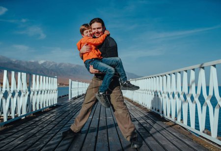 Foto de Feliz padre sostiene a su hijo en sus brazos de pie en un muelle en el fondo de las montañas - Imagen libre de derechos