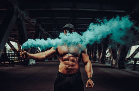 Foto de Joven hombre fuerte culturista de pie con denso humo azul en el fondo industrial urbano - Imagen libre de derechos