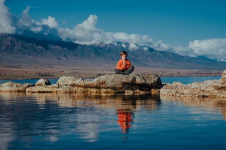 Boy sitting in meditation pose on rock in lake on mountains background