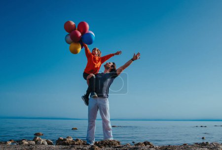 Foto de Father holding his son with balloons on shore of the lake on summer day - Imagen libre de derechos