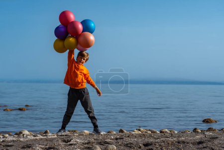 Foto de Cheerful boy standing with balloons on lake shore on summer day - Imagen libre de derechos