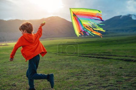 Foto de Happy boy runs with kite on green field on mountains background at sunset - Imagen libre de derechos