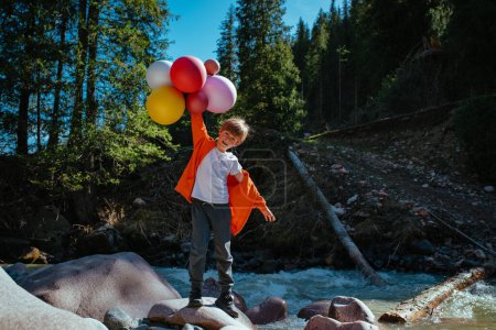 Téléchargez les photos : Garçon heureux avec des ballons se dresse sur la rive de la rivière de montagne - en image libre de droit
